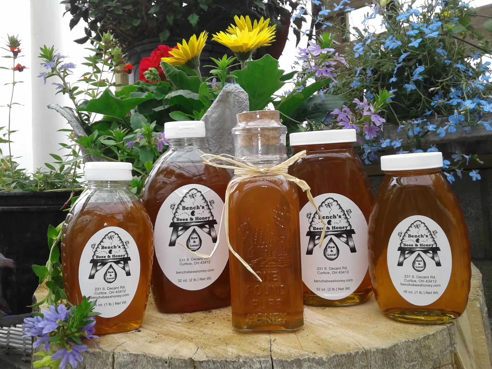 Five glass and plastic jars of Bench's Bees & Honey raw honey, labeled and tied with raffia, displayed among red, yellow, and blue garden flowers
